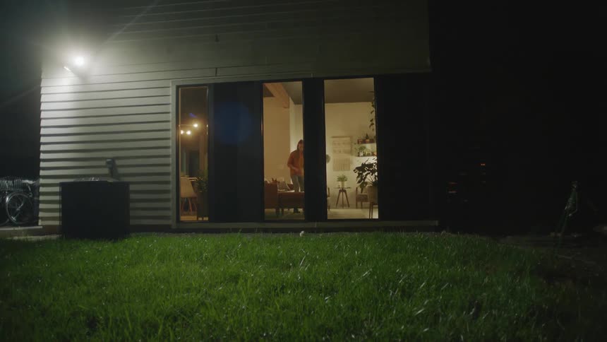 Exterior of backyard looking into a living room as a woman sits down on the couch in the living room