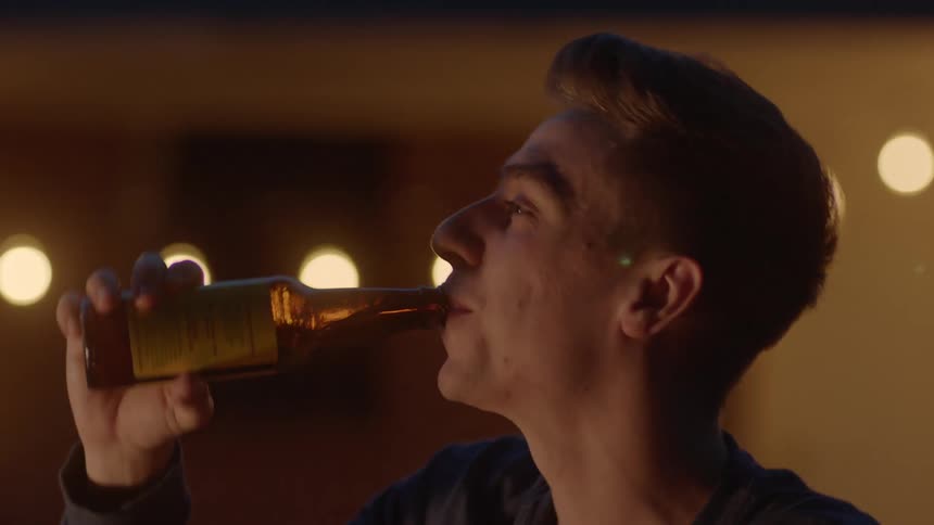 A smiling young man drinks liquor while sitting outdoors at night