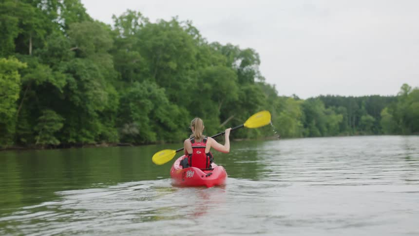 Young blonde woman paddling on a red kayak on a river through the forest
