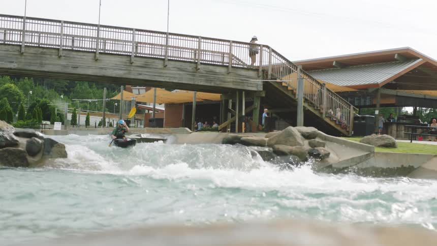Man kayaking at daytime in a river within a park under a bridge while a woman walks overhead