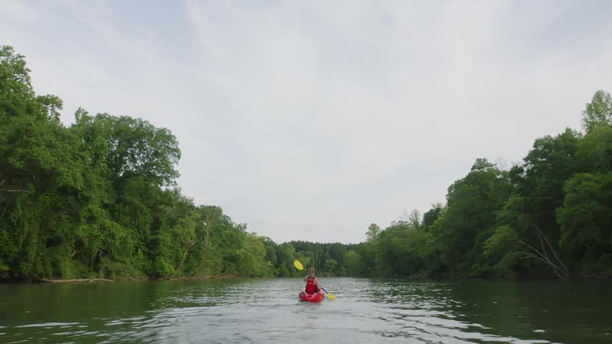 A woman paddles alone in a red kayak down the center of a river