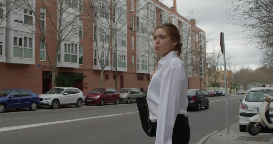 Blue car passes in front of woman wearing a white blouse and black pants standing on the sidewalk