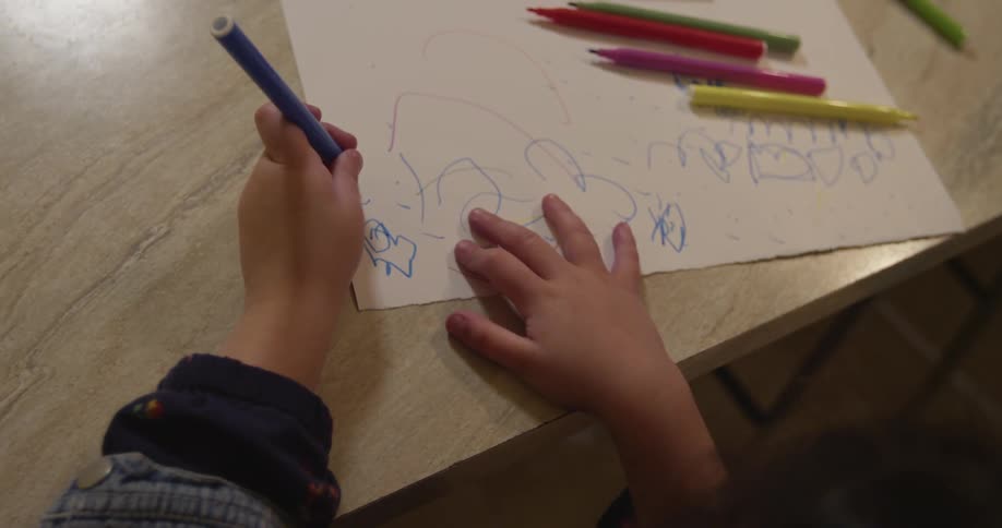 A child sits at a table drawing with a blue colored pencil on a piece of paper indoors