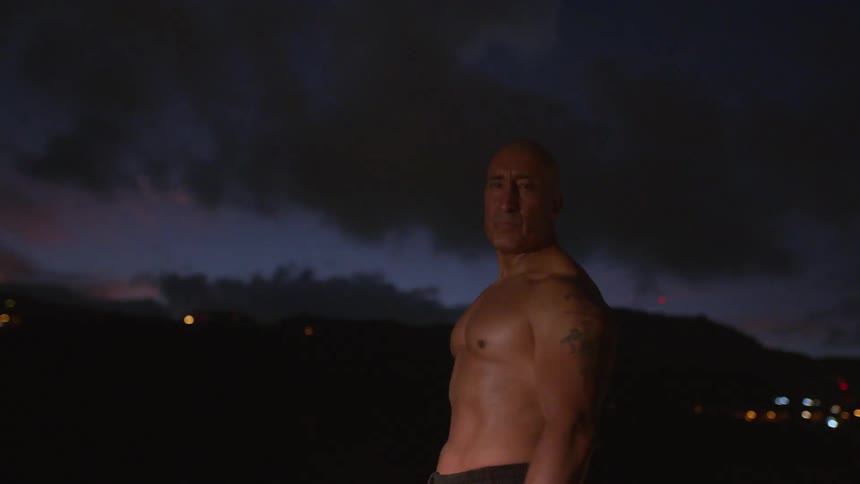 A man with large muscles walks in front of a city skyline without a shirt on