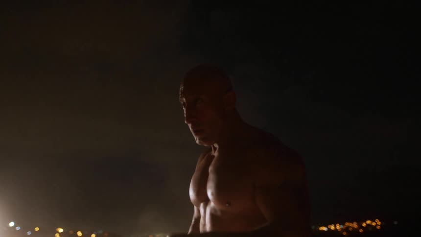 A large muscular man practices throwing punches in the darkness of night