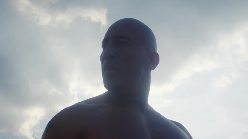 A shirtless man stands outside and looks over his shoulder