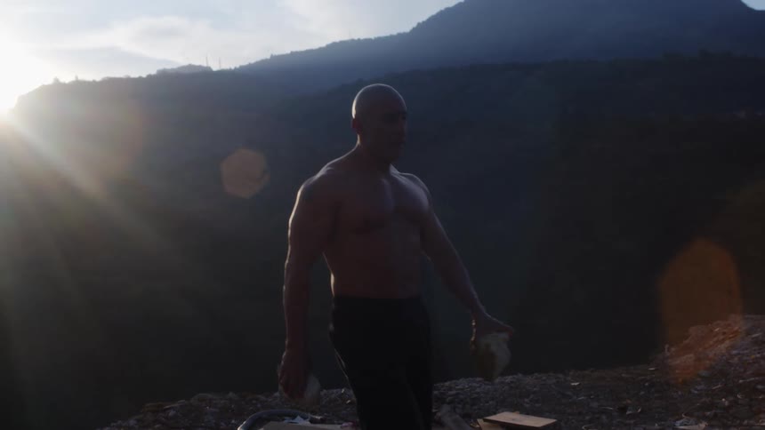 Shirtless muscular bald man carries rocks near the edge of a cliff in the mountains at sunset