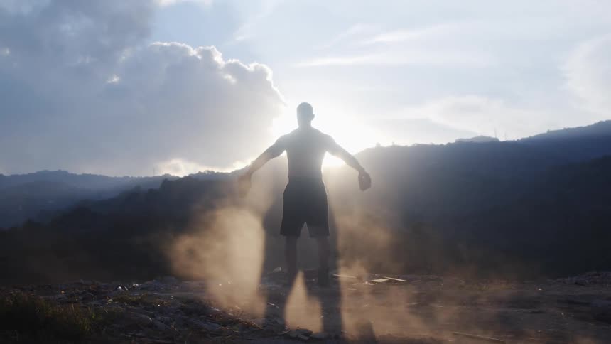 Sunlight shines over a distant mountain onto a shirtless man lifting stones on a cliff while facing the rising sun
