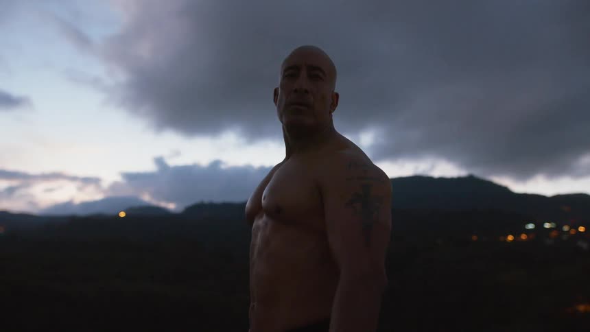 A large muscular man with an shaved head stands outside at dusk