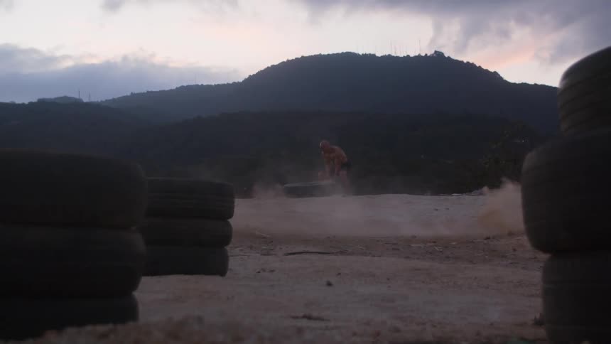 A muscular shirtless man flips a large tire end over end in a dusty flatland on a cloudy day