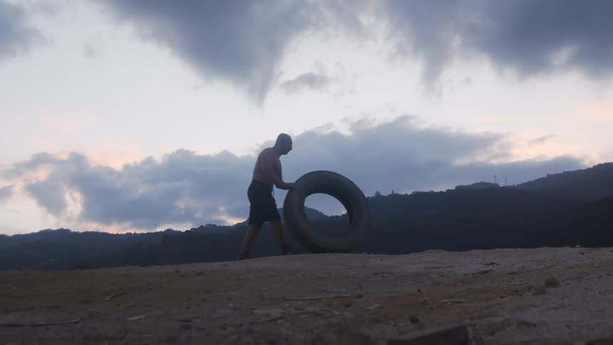 Shirtless man in shorts carefully rolls a large tire across bare dirt in front of a background of hills and clouds
