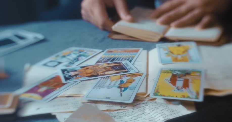 Mans hands flipping through a small book on a desk and arranging tarot cards in a stack