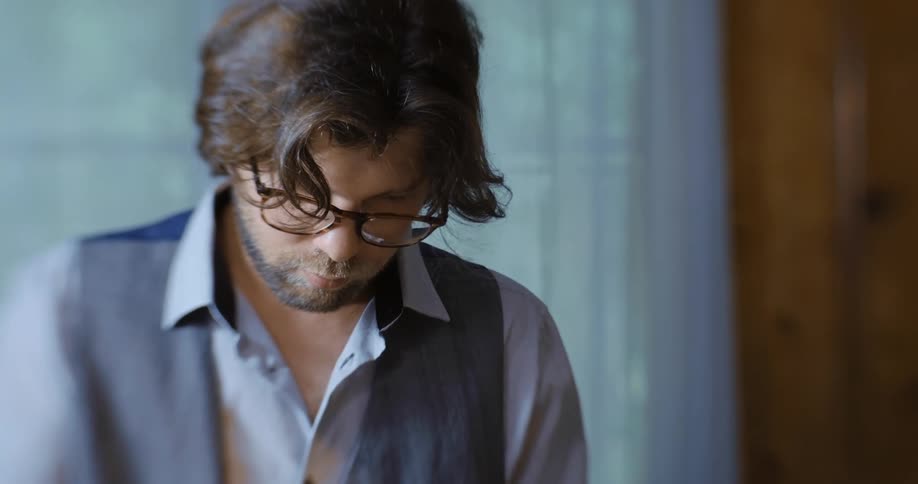 A man wearing glasses and a blue dress shirt and vest stands inside looking at photos