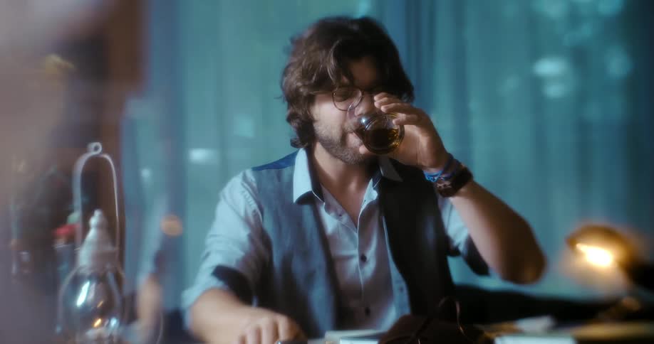 A man sits and his desk and takes a drink of a dark liquor