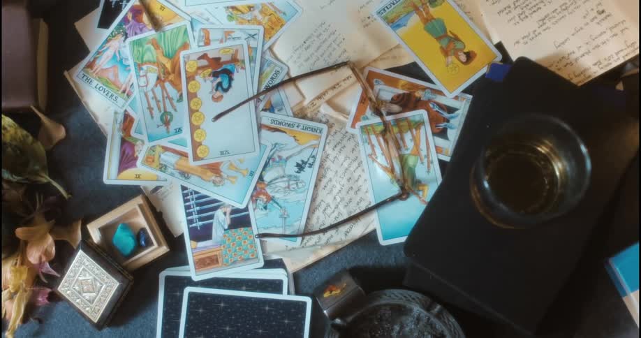 A pair of eyeglasses is dropped onto a desk covered with tarot cards papers and a glass with liquor