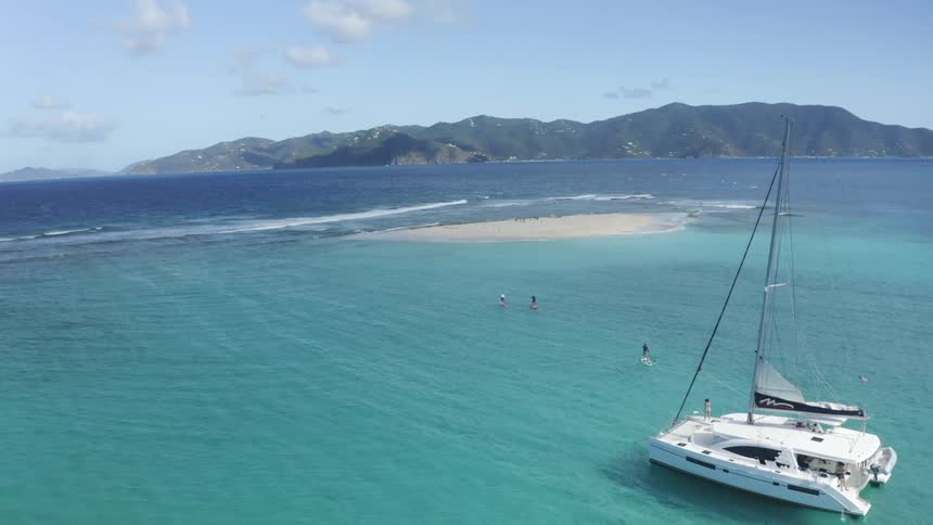 A group of paddle boarders paddle away from a yacht towards a sand bar in the light blue waters of the ocean during the day