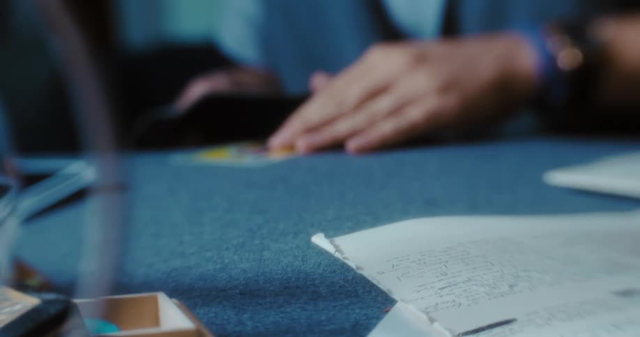 A man with a beard and glasses gathers cards in his hands as he sits at a table with paper and a glass
