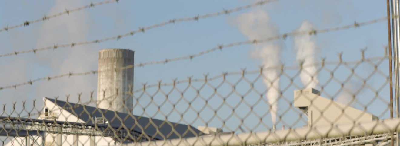 Smoke coming from exhaust pipes on a factory roof top from behind a fence