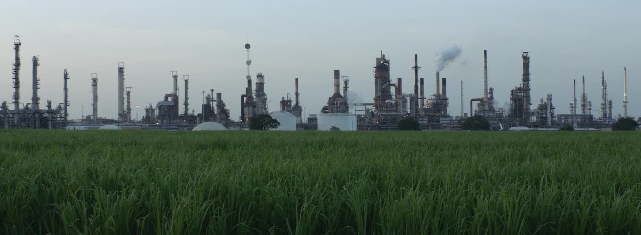 Across a wide open grass field a large refinery facility can be seen