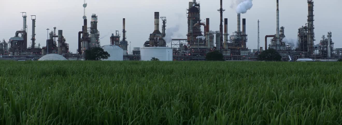 Smoke billows from a large refinery in a grassland on a cloudy day
