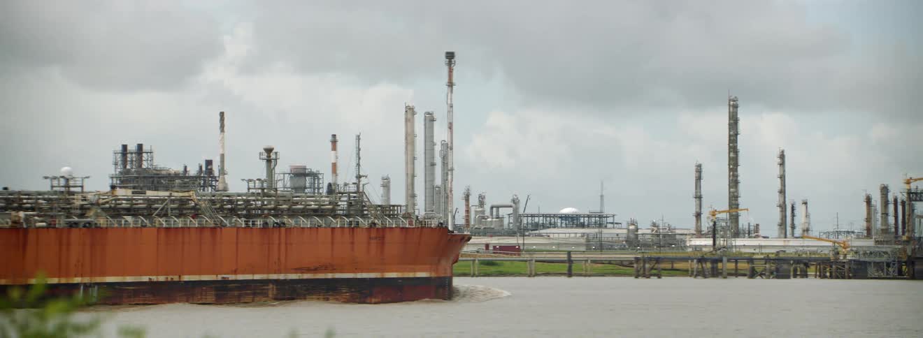A tanker ship moving on a river with a factory the sky and clouds in the background