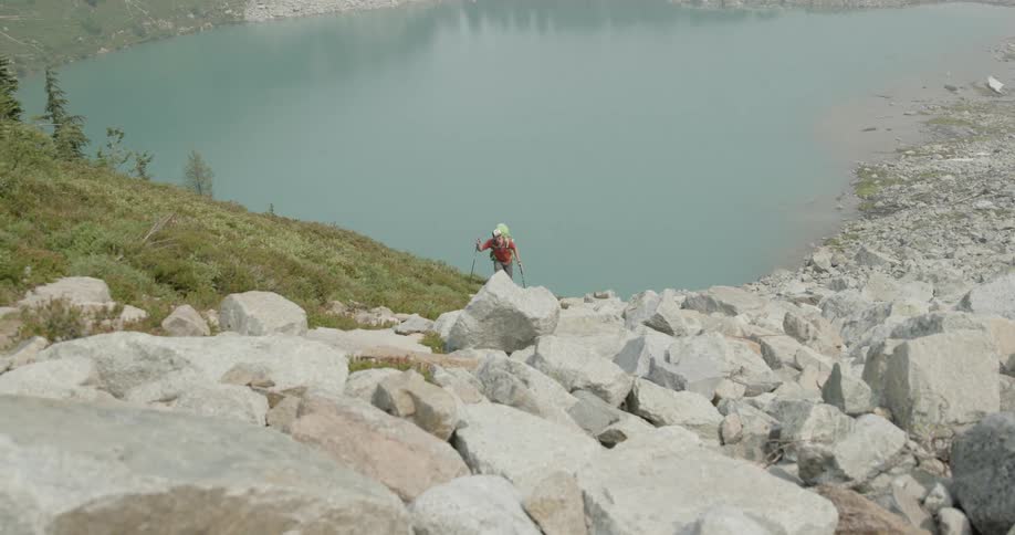A man hikes up a steep hill with the help with poles with a lake behind him and rocks on the left
