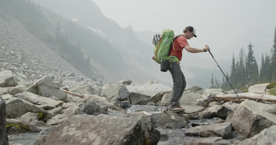 A man walks over a stream and rocks by a mountain during the day