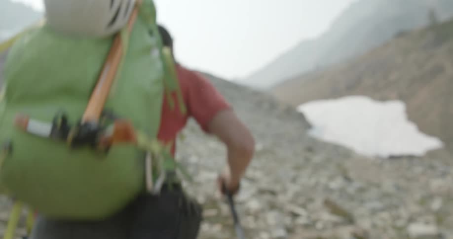 A man hikes along a rocky mountain terrain with walking sticks and a large backpack