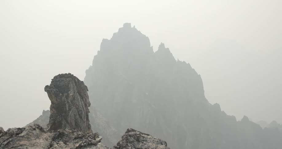 A jagged and rocky mountain peak stands tall as it is surrounded by a foggy haze