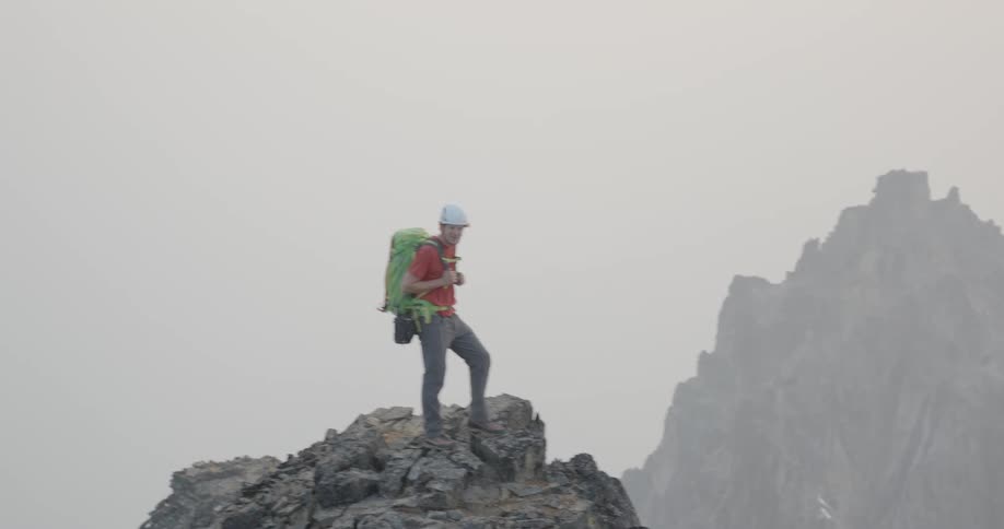 A man with a helmet and a backpack standing on a cliff with the sky and clouds in the background
