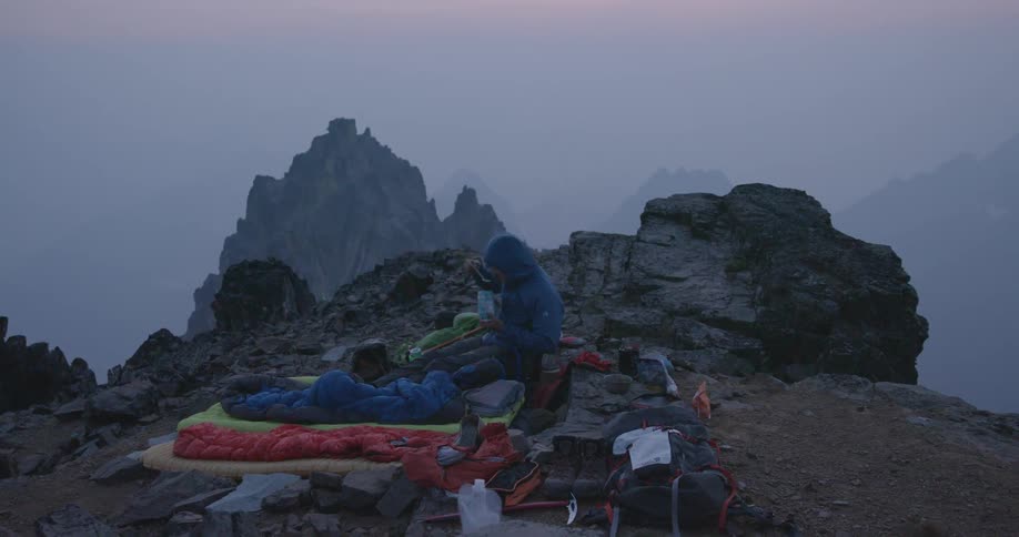 Person wearing coat sits in camp eating on mountain with fog obscuring other mountain peaks