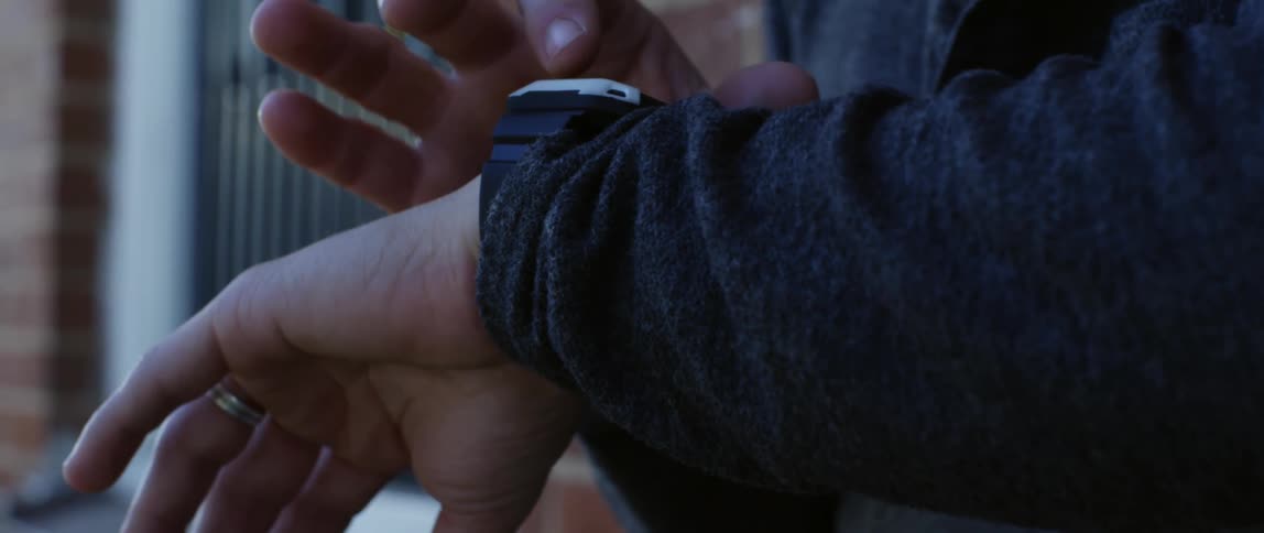 A man with tan skin is standing next to a red brick structure wearing a long sleeved gray shirt playing with his watch