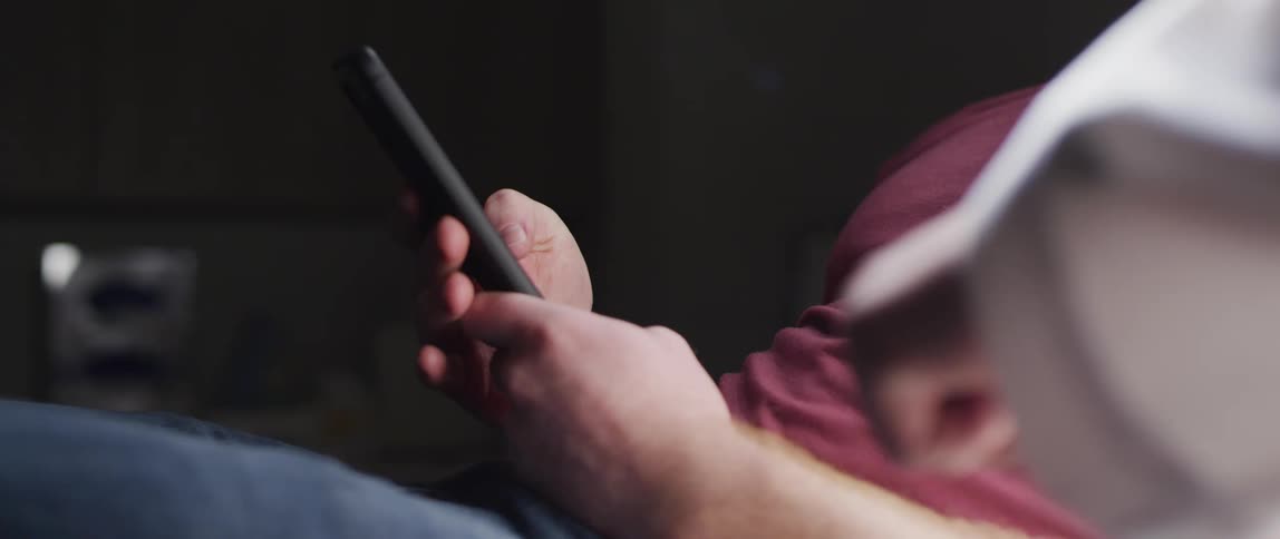 A man is sitting sitting on a sofa looking at his smartphone while another person is pointing their fist towards him in the foreground