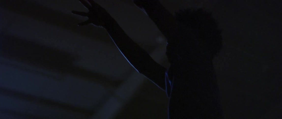 A young male stands inside of a darkened gymnasium where he shoots a basketball