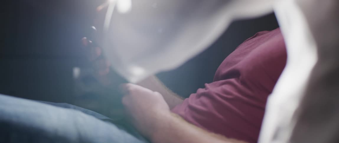 Man wearing red shirt resting on a bed using a smartphone in a dimly lit room