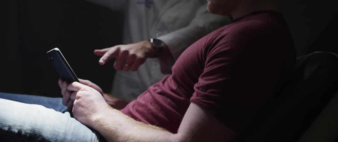 Doctor in white coat reaches over to a man on a smart phone and points at his screen with a light shining on him