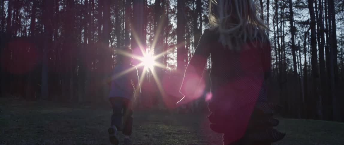 A boy and a girl playfully running towards the treeline at sunset