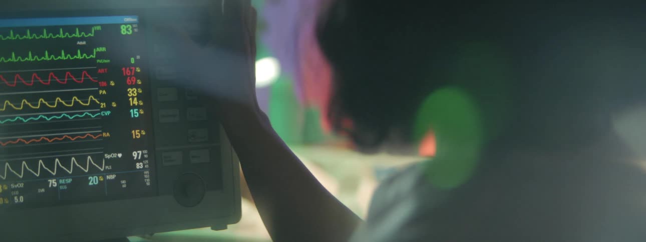 A nurse in the hospital touching buttons on a monitor with a person laying in a hospital bed in the background