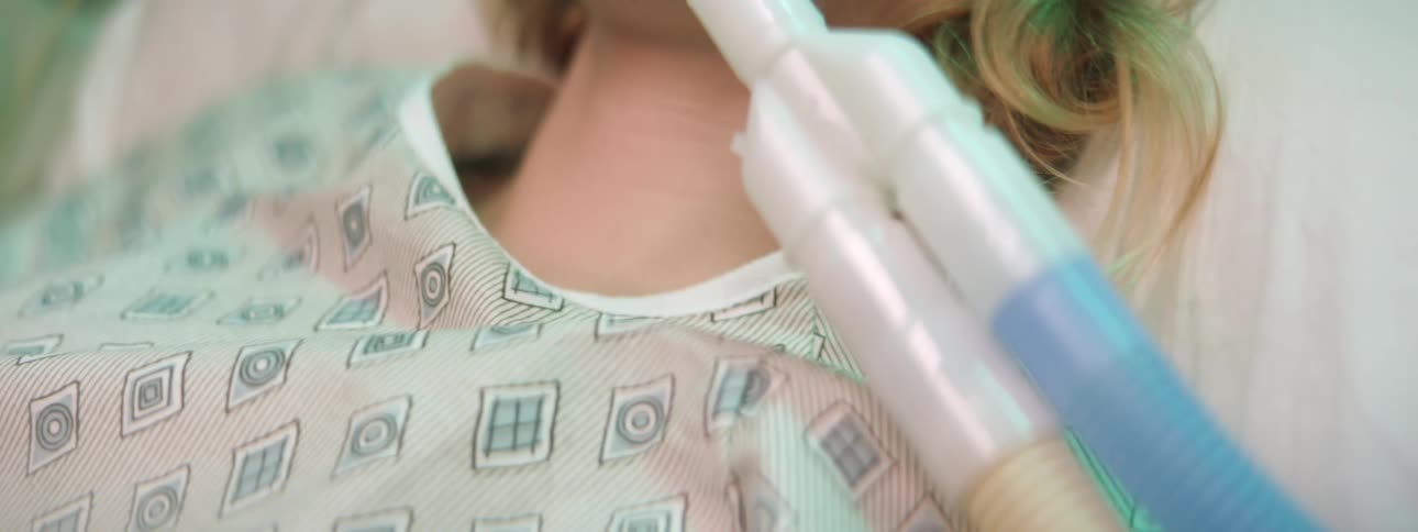A blonde woman is on a ventilator lying on a hospital bed wearing a hospital gown