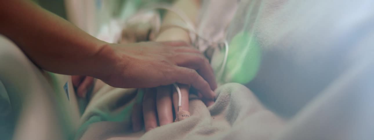 A person sitting on the side of a hospital bed grips the hand of the resting patient