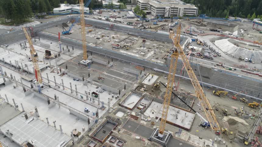 Construction is underway on a large building foundation covered with cranes and heavy machinery