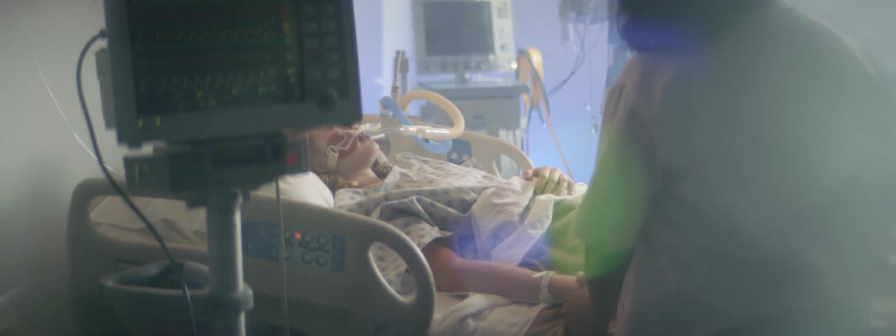 A woman holds her daughters hand as she lies in a hospital bed hooked up to tubes