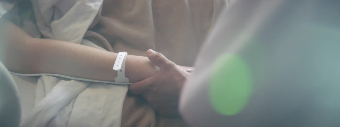 A person holds the hand of a person wearing a hospital bracelet and lying in a hospital bed