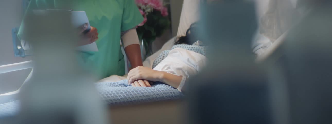 A woman lies in a hospital bed while a medical staff member offers comfort