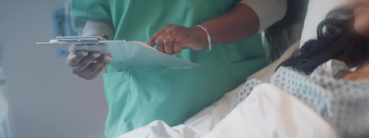Female patient lying in a hospital bed while a nurse stands by reading from a clipbaord
