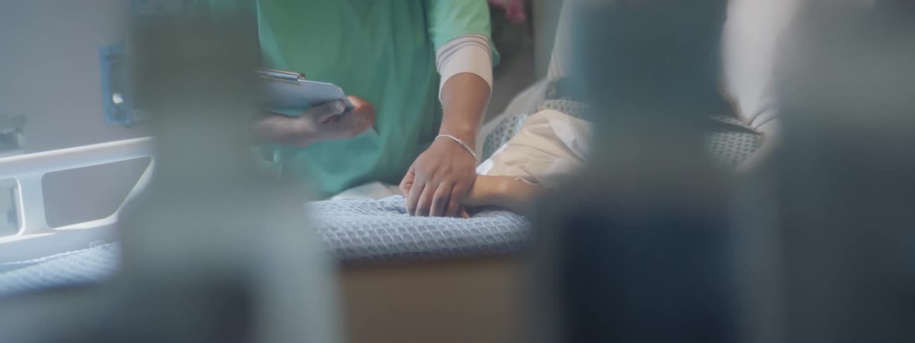 A nurse places her hand on the hands of a patient in a hospital bed