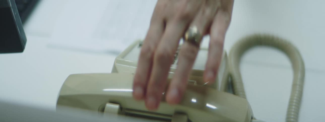 A woman sitting at a desk picks up the receiver of a cream colored phone