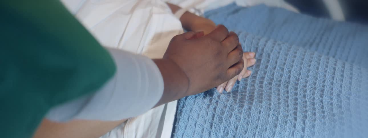 A woman lies in a hospital bed while a medical employee looks at a chart and holds her hands