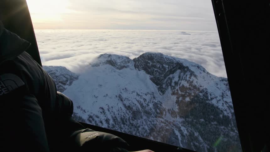 A person sitting with a hat and jacket with a mountain the sky and clouds out the window
