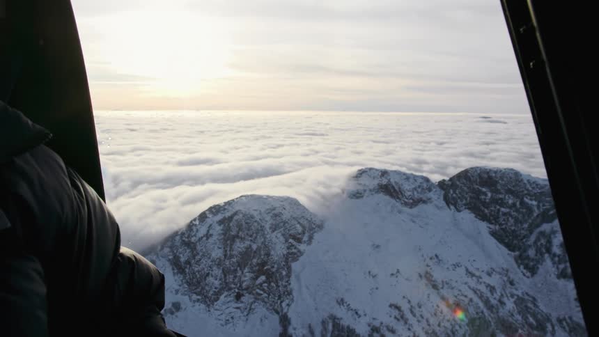 A man sits inside an airplane looking out the window to mountains cloud cover and a low sun